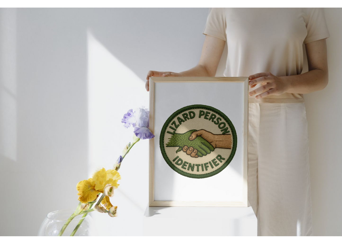 Person holding a framed picture with a 'Lizard Person Identifier' design, next to flowers on a white surface.