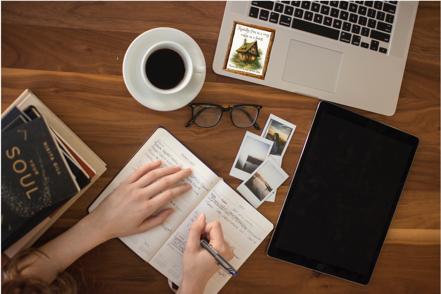 Person writing in a notebook on a wooden desk with a laptop, tablet, coffee, and books.