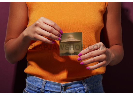 Person holding a card with an alien UFO image against an orange shirt.